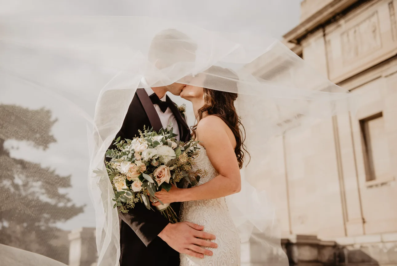 Jeunes mariés s'embrassant sous un voile de mariée, bouquet de fleurs au premier plan